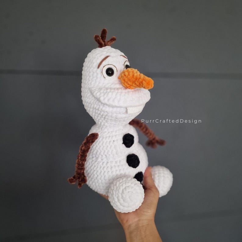 May include: A handmade white crochet snowman with a carrot nose, black button details, and brown twig arms and hair. The snowman is a character from the movie Frozen. The text "PurrCraftedDesign" is visible.