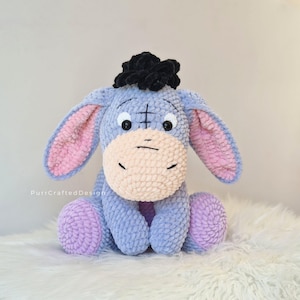 Pode incluir: Um brinquedo de pel&uacute;cia feito &agrave; m&atilde;o de um burro, com corpo azul claro, orelhas internas rosa e focinho bege. O burro tem um tufo de cabelo preto e est&aacute; sentado com as pernas esticadas. O brinquedo &eacute; feito de material macio e texturizado.