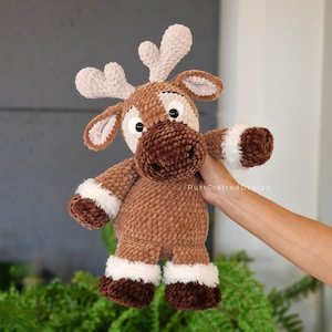 May include: A plush brown and white reindeer toy with large, expressive eyes and antlers. The reindeer has a soft, textured body, white accents on its hooves and arms, and a dark brown snout. The toy is handmade and appears to be a children's item.