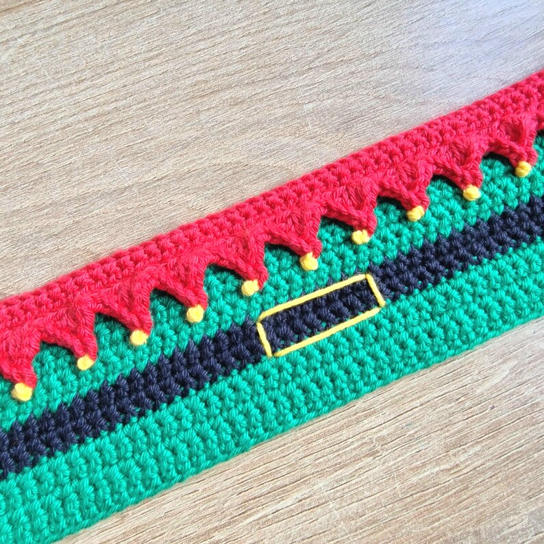 May include: A close-up of a green, black, and red crocheted trim with yellow accents. The trim features a black belt with a yellow outline.