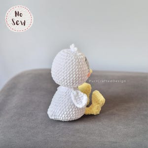 K&ouml;nnte beinhalten: Ein handgefertigtes wei&szlig;es H&auml;kelk&uuml;ken mit gelben F&uuml;&szlig;en und einem grauen Schnabel. Das K&uuml;ken sitzt auf einer grauen Oberfl&auml;che. Der Text "No Sew" befindet sich in einem Kreis oben links.