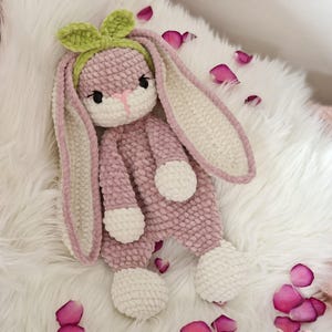 May include: A crocheted pink bunny rabbit with white accents and a green bow. The bunny is sitting on a white fluffy surface with pink rose petals.