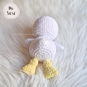 K&ouml;nnte beinhalten: Eine handgefertigte wei&szlig;e H&auml;kelente mit gelben F&uuml;&szlig;en. Die Ente liegt auf einer wei&szlig;en, flauschigen Oberfl&auml;che. Der Text "No Sew" befindet sich in einem Kreis oben links. Der Text "PurrCraftedDesign" befindet sich unten links.