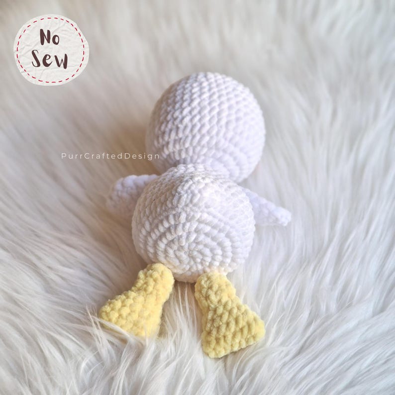 K&ouml;nnte beinhalten: Eine handgefertigte wei&szlig;e H&auml;kelente mit gelben F&uuml;&szlig;en. Die Ente liegt auf einer wei&szlig;en, flauschigen Oberfl&auml;che. Der Text "No Sew" befindet sich in einem Kreis oben links. Der Text "PurrCraftedDesign" befindet sich unten links.
