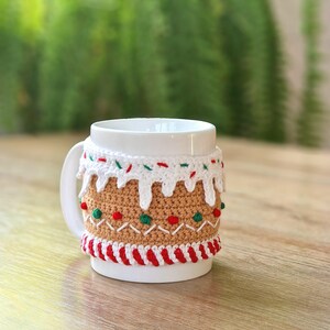 May include: White ceramic mug with a crocheted gingerbread house design. The design features red and green sprinkles, white icing, and a brown gingerbread base.