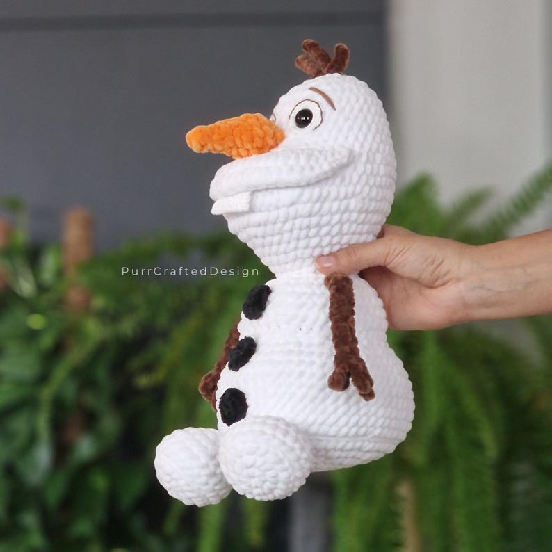 May include: A white crocheted Olaf doll with a carrot nose, brown twig hair, and black button accents. The doll has a smiling expression and is holding a pose. The text "PurrCraftedDesign" is visible on the lower left.