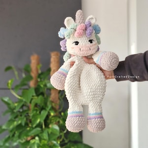 Puede incluir: Un unicornio de peluche blanco tejido a crochet con una cara rosa y una melena de colores pastel. El unicornio está sosteniendo una manta en sus pezuñas.