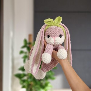 May include: A pink and white crocheted bunny with long ears and a green headband. The bunny is holding its paws together and has a sweet expression.