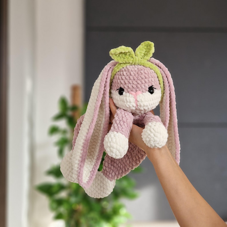 May include: A pink and white crocheted bunny with long ears and a green headband. The bunny is holding its paws together and has a sweet expression.