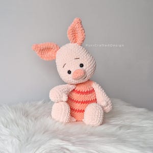 May include: A handmade plush piglet toy, predominantly light pink with peach detailing. The piglet has large, floppy ears, a small snout, and black eyes. The toy is seated on a white, fluffy surface, creating a soft texture.