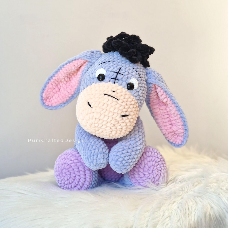 Pode incluir: Um brinquedo de pel&uacute;cia artesanal de burro em tons de azul, roxo e rosa. O burro tem orelhas grandes com o interior rosa, um focinho bege claro e um tufo de cabelo preto. O brinquedo est&aacute; sentado em uma superf&iacute;cie branca e fofa.