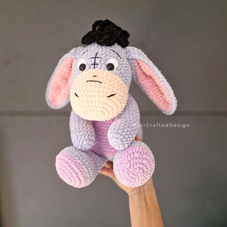 Pode incluir: Um brinquedo de pel&uacute;cia feito &agrave; m&atilde;o do Eeyore, o burro de Winnie the Pooh. A pel&uacute;cia &eacute; principalmente lavanda com detalhes em rosa nas orelhas e nos p&eacute;s. Possui um focinho creme, olhos pretos e um pequeno tufo de cabelo preto.