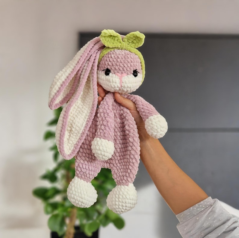 May include: A pink and white crocheted bunny rabbit plush toy with a green bow. The bunny has a white face with black eyes and a white nose.
