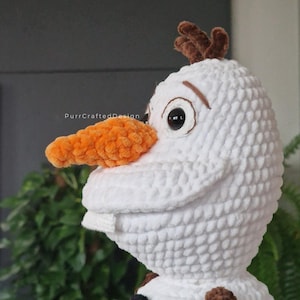 May include: A handmade white crochet Olaf doll with an orange carrot nose, black button eyes, and brown twig hair. The doll has a smiling expression and is a soft, plush toy. The text "PurrCraftedDesign" is visible.
