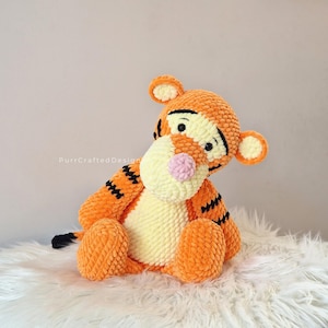 Puede incluir: Un peluche de tigre naranja con rayas negras, detalles amarillos y una nariz rosa. El animal de peluche hecho a mano está sentado sobre una superficie blanca y esponjosa. El tigre tiene orejas pequeñas y una cola negra. Juguete para niños.