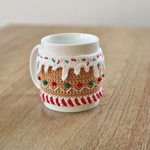 May include: White ceramic mug with a crocheted gingerbread house design. The design features red and green candy sprinkles, white icing, and brown gingerbread.