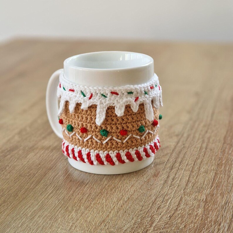 May include: White ceramic mug with a crocheted gingerbread house design. The design features red and green candy sprinkles, white icing, and brown gingerbread.