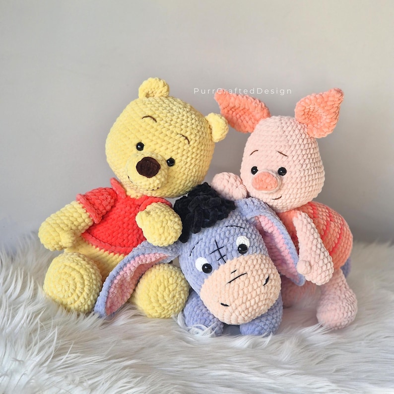 Pode incluir: Pel&uacute;cias de croch&ecirc; feitas &agrave; m&atilde;o de Ursinho Pooh, Leit&atilde;o e Eeyore. O urso amarelo usa uma camisa vermelha, o porco rosa usa uma camisa vermelha e rosa, e o burro azul tem um tufo de cabelo preto. Os brinquedos est&atilde;o em uma superf&iacute;cie branca e fofa.