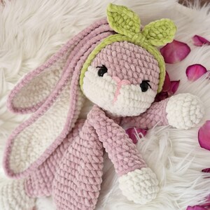 May include: A pink crocheted bunny with white accents and a green crocheted headband. The bunny is lying on a white fluffy surface.