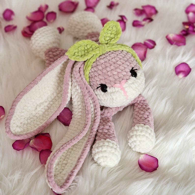 May include: A pink and white crocheted bunny with long ears and a green bow. The bunny is lying on a white fluffy surface with pink rose petals scattered around it.
