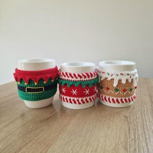 May include: Three white ceramic mugs with crocheted Christmas-themed cozies. The cozies are red, green, and white with a gingerbread man, a candy cane, and an elf design.