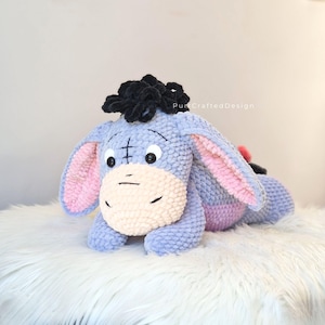 Pode incluir: Um brinquedo de pel&uacute;cia artesanal do Eeyore, o burro de Winnie the Pooh. A pel&uacute;cia &eacute; azul clara com o interior das orelhas rosa, um focinho creme e uma crina preta. O brinquedo est&aacute; deitado sobre uma superf&iacute;cie branca e fofa. O texto "Pun Crafted Design" &eacute; vis&iacute;vel.