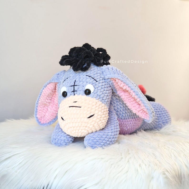 Pode incluir: Um brinquedo de pel&uacute;cia artesanal do Eeyore, o burro de Winnie the Pooh. A pel&uacute;cia &eacute; azul clara com o interior das orelhas rosa, um focinho creme e uma crina preta. O brinquedo est&aacute; deitado sobre uma superf&iacute;cie branca e fofa. O texto "Pun Crafted Design" &eacute; vis&iacute;vel.