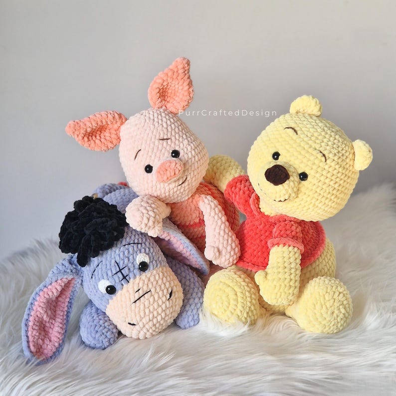 Pode incluir: Tr&ecirc;s brinquedos de pel&uacute;cia de croch&ecirc; feitos &agrave; m&atilde;o: um urso amarelo com uma camisa vermelha, um porco rosa e um burro azul. Os brinquedos s&atilde;o macios e fofinhos, perfeitos para crian&ccedil;as ou colecionadores. Os brinquedos est&atilde;o dispostos em uma superf&iacute;cie branca e fofa.