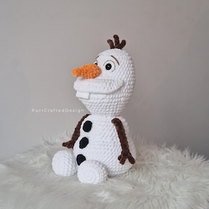 May include: A handmade crochet Olaf doll, featuring a white body, an orange carrot nose, and black button accents. The doll has brown twig-like hair and arms, and a friendly smile. The doll is sitting on a fluffy white surface.