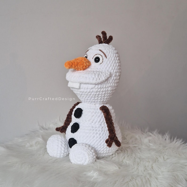 May include: A handmade crochet Olaf doll, featuring a white body, an orange carrot nose, and black button accents. The doll has brown twig-like hair and arms, and a friendly smile. The doll is sitting on a fluffy white surface.
