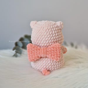 Puede incluir: Un oso de ganchillo rosa claro hecho a mano con un lazo coral. El oso tiene orejas pequeñas y una cola diminuta. El lazo es de un color coral contrastante. El oso está sobre una superficie blanca y esponjosa.
