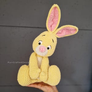 May include: A handmade yellow crochet bunny with pink inner ears, a pink nose, and black eyes. The plush toy is sitting with its arms folded and is made with a textured yarn. The bunny is a soft toy.