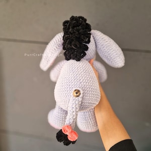 Pode incluir: Um brinquedo de pel&uacute;cia de burro de croch&ecirc; roxo claro feito &agrave; m&atilde;o com uma crina e cauda pretas e uma ponta de cauda coral e preta. O burro tem um bot&atilde;o na parte de tr&aacute;s e o texto "PurrCraftDesign" &eacute; vis&iacute;vel.