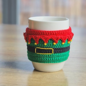 May include: White ceramic mug with a crocheted elf-themed cozy. The cozy is green, black, and red with yellow accents.