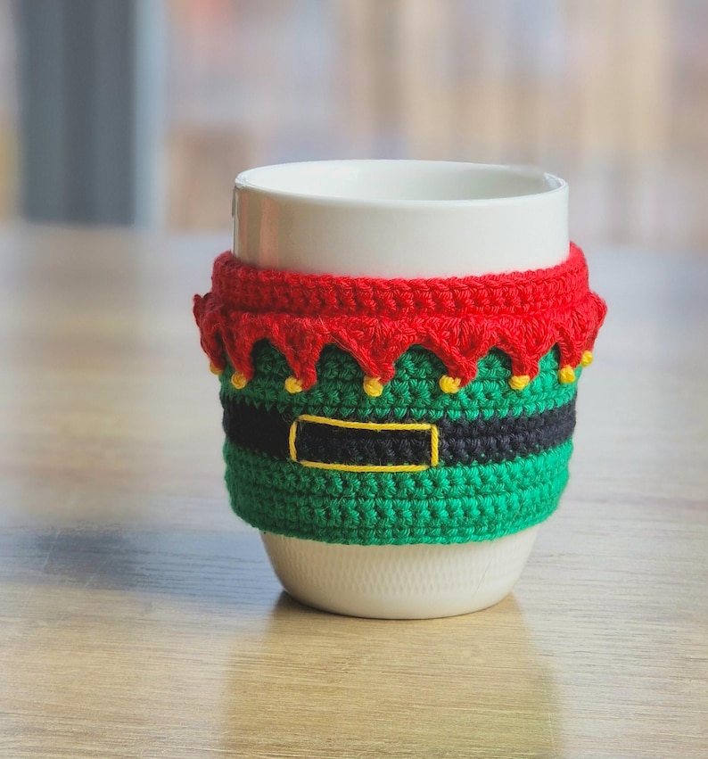 May include: White ceramic mug with a crocheted elf-themed cozy. The cozy is green, black, and red with yellow accents.