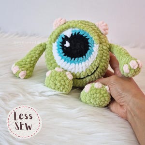 Puede incluir: Un peluche verde tejido a ganchillo con un ojo grande y redondo con detalles azules, blancos y negros. El juguete tiene brazos y pies pequeños con detalles rosas. El texto "Less Sew" está en un círculo en la parte inferior.