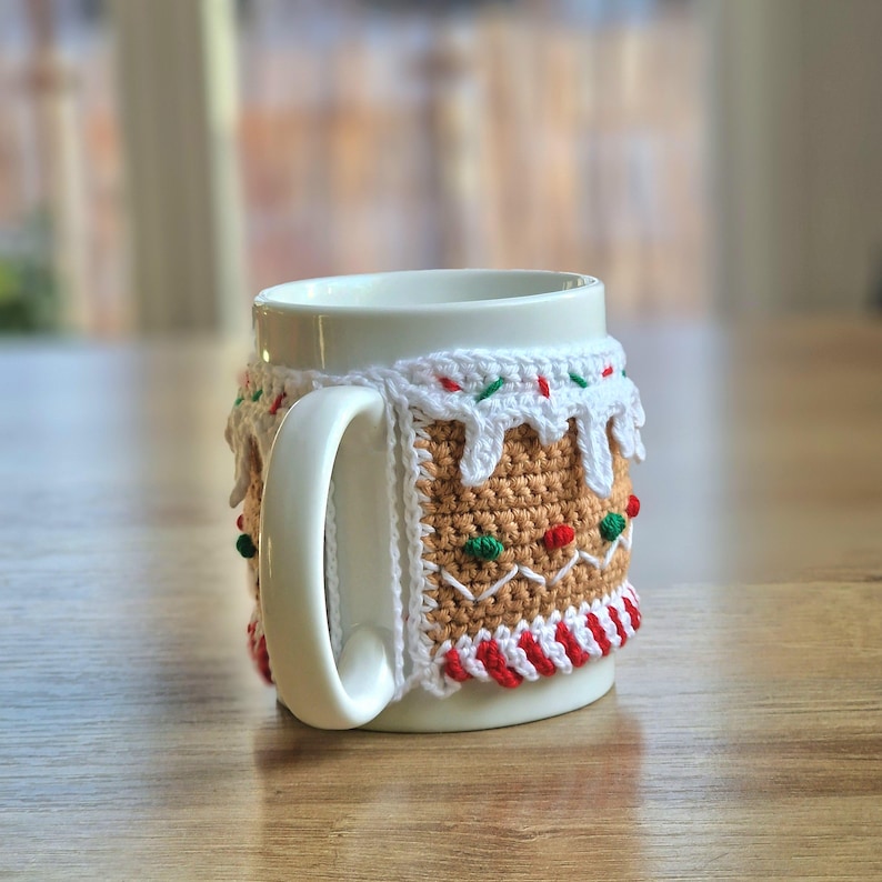 May include: White ceramic mug with a crocheted gingerbread house cozy. The cozy is brown, white, red, and green and features candy cane stripes and white icing.
