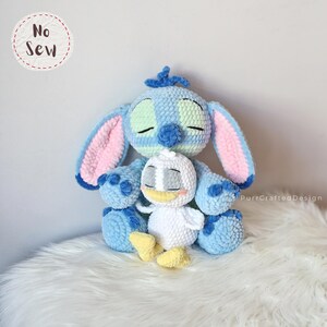 K&ouml;nnte beinhalten: Ein handgefertigtes blau-wei&szlig;es Stitch-Pl&uuml;schtier aus H&auml;kelarbeit mit einem kleineren wei&szlig;en Enten-Pl&uuml;schtier. Das Stitch-Pl&uuml;schtier hat gro&szlig;e Ohren mit rosa Akzenten, und die Ente hat einen gelben Schnabel und F&uuml;&szlig;e. Die Spielzeuge liegen auf einer wei&szlig;en, flauschigen Oberfl&auml;che.