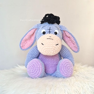 Pode incluir: Um brinquedo de pel&uacute;cia artesanal do Eeyore, o personagem burro de Winnie the Pooh. A pel&uacute;cia &eacute; principalmente azul com orelhas internas rosa, um focinho bege claro e p&eacute;s roxos. Tem um tufo de cabelo preto e est&aacute; sentado em uma superf&iacute;cie branca.