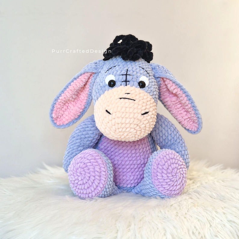 Pode incluir: Um brinquedo de pel&uacute;cia artesanal do Eeyore, o personagem burro de Winnie the Pooh. A pel&uacute;cia &eacute; principalmente azul com orelhas internas rosa, um focinho bege claro e p&eacute;s roxos. Tem um tufo de cabelo preto e est&aacute; sentado em uma superf&iacute;cie branca.