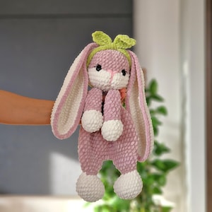 May include: A crocheted pink bunny with white accents and a green bow. The bunny has long ears and a fluffy texture.