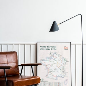 May include: A black and brown leather armchair with a wooden armrest sits in front of a framed map of France titled "Carte de France du voyage &agrave; v&eacute;lo". A black floor lamp with a conical shade is in the background.