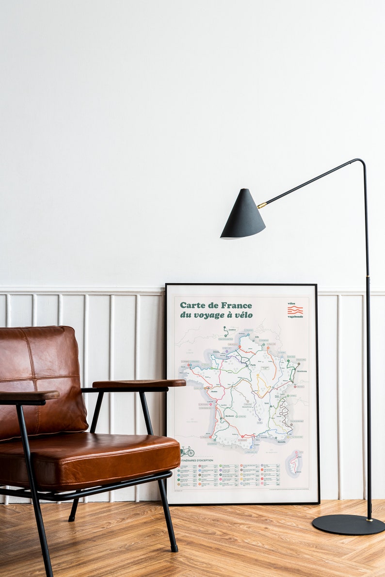 May include: A black and brown leather armchair with a wooden armrest sits in front of a framed map of France titled "Carte de France du voyage &agrave; v&eacute;lo". A black floor lamp with a conical shade is in the background.