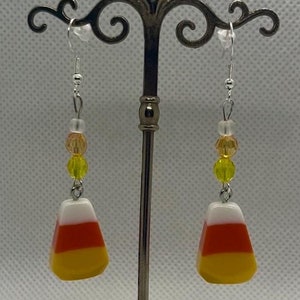 May include: A pair of dangle earrings featuring candy corn-shaped charms with white, orange, and yellow stripes. The charms are accented with clear, yellow, and orange beads.