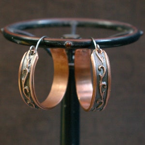 May include: A pair of copper hoop earrings with a dark, ornate design. The earrings are suspended from a dark metal structure, showcasing their intricate details and warm, metallic tones. The design features a dark, swirling pattern.