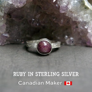 May include: A sterling silver ring featuring a round, cabochon-cut ruby gemstone. The silver band displays a repeating chevron pattern. Text at the bottom reads: RUBY IN STERLING SILVER, Canadian Maker, accompanied by a Canadian flag icon.