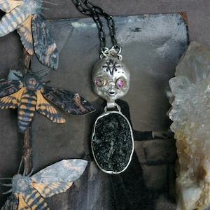May include: A silver-toned pendant necklace featuring a stylized skull with iridescent eyes and a black symbol on the forehead, suspended from a dark chain. Below the skull is an oval frame containing a textured, dark, crystalline mineral stone.