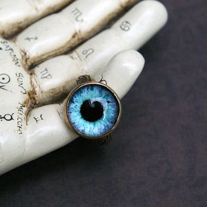 May include: A round pendant featuring a detailed blue eye design with a black pupil, set in a gold-colored metal frame. The pendant is displayed on a white, textured surface with astrological symbols.
