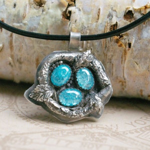 Könnte beinhalten: Eine silberne Anhängerhalskette mit drei ovalen, leuchtend blauen Edelsteinen. Der Anhänger hat ein strukturiertes, organisches Design mit Blattakzenten und hängt an einer schwarzen Kordel.