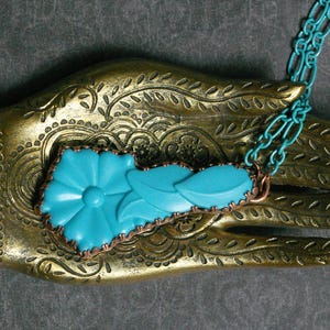 May include: A turquoise floral pendant necklace with a copper border and a matching turquoise chain. The pendant features a flower design with leaves. The necklace is displayed on a gold, ornate hand.